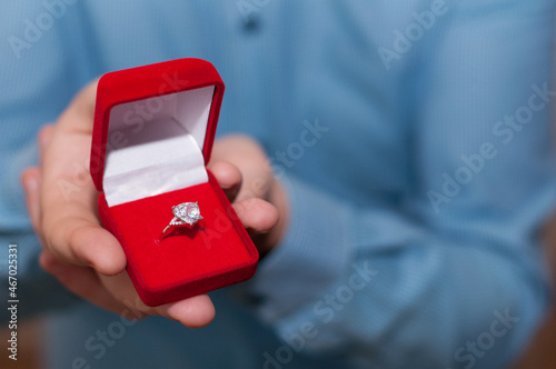 A white gold ring with a precious stone in an open red velvet box in the hand of a man in a blue shirt