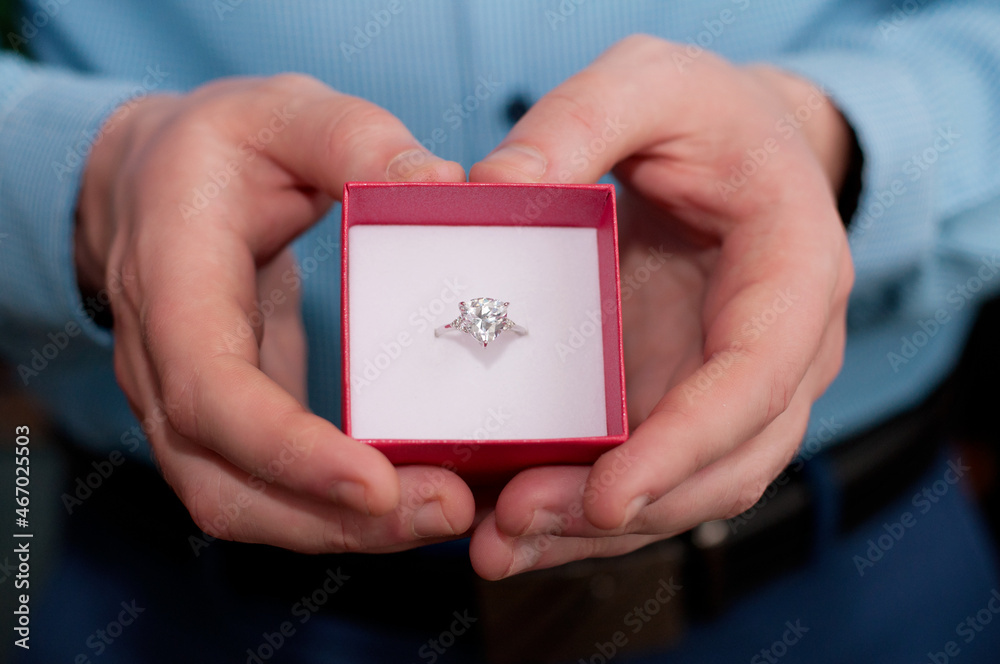 A red box with a white gold ring and a precious stone in the heart from the hands of a man in a blue shirt A red box with a white gold ring and a precious stone in the heart from the hands of a man in a blue shirt