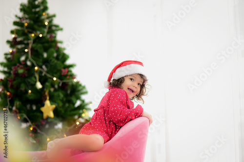 A cute girl in a red polka dot dress and wearing Santa Claus hat bright and cheerful are playing on the sofa and behind it there is a Christmas tree with twinkling lights.