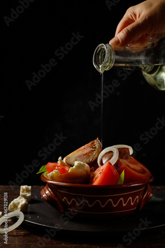 Homemade salad of different varieties of tomatoes, onions and homemade soft cheese in a clay bowl. And the hand pours oil from a glass jug. Low key, Vertical, Selective focus