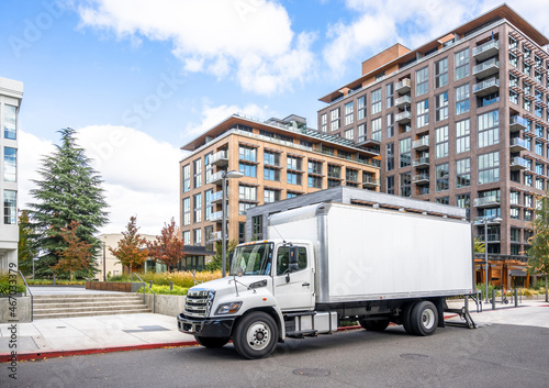Middle size rig white semi truck with box trailer unloading cargo delivered to new multilevel apartment buildings in urban city