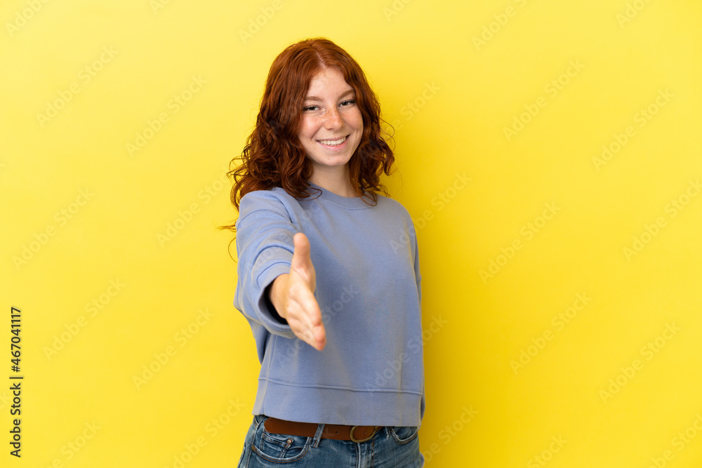 Teenager reddish woman isolated on yellow background shaking hands for closing a good deal Teenager reddish woman isolated on yellow background shaking hands for closing a good deal