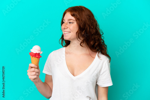Teenager reddish woman with a cornet ice cream isolated on blue background looking to the side and smiling