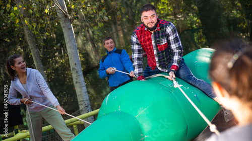 Emotional bearded young man sitting astride big inflatable rodeo bottle, having fun with friends in outdoor amusement park