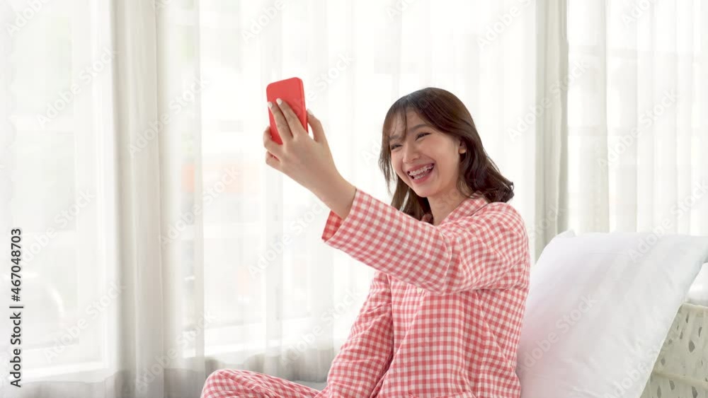 A beautiful Asian woman in red pajamas sits in bed using her smart phone to video call her boyfriend or friend to say hello in the morning.
