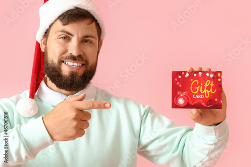 Handsome man in Santa hat pointing at gift card on pink background photo
