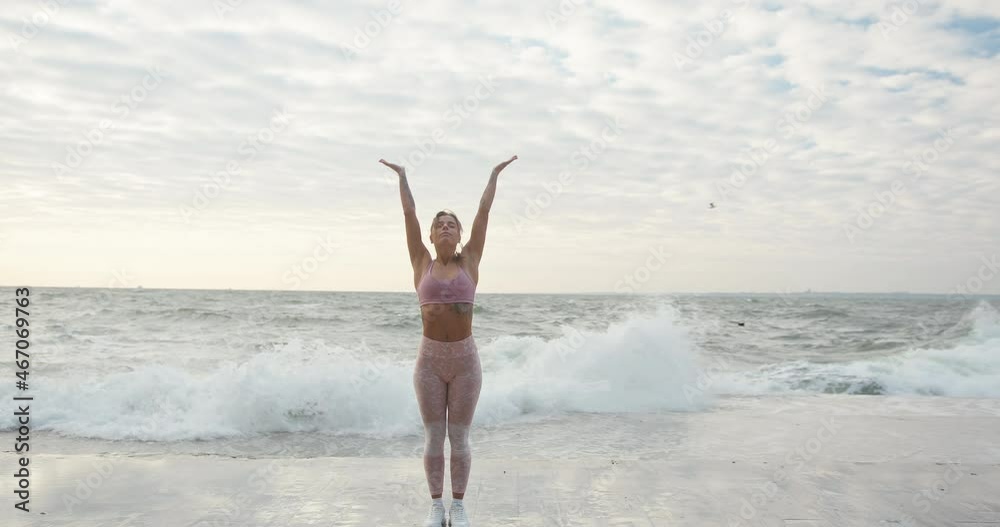 Blonde-haired woman with ponytail in pink top and leggings does yoga and meditates on embankment against stormy sea waves slow motion Blonde-haired woman with ponytail in pink top and leggings does yoga and meditates on embankment against stormy sea waves slow motion