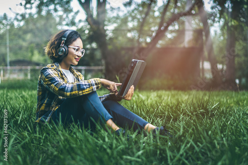 Asian hipster curly girl sitting on the lawn She wears headphones and smiles happily while using the tablet to relax and study online. in the atmosphere of the soft sunlight in the morning. © brain2hands