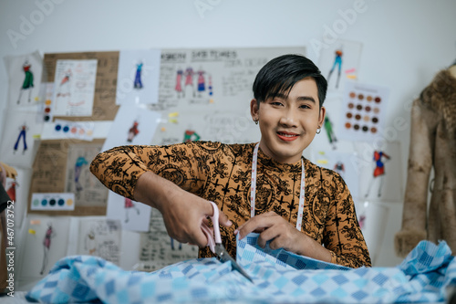 Portrait of Asian young male tailor cutting piece of cloth at table in fashion studio. Designer and LGBTQ Concept.