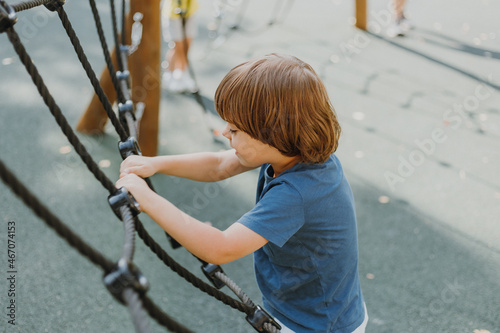 pretty little brunette boy in a blue T-shirt is climbing a rope web. child is engaged in outdoor sports. healthy active kid. lifestyle. space for text. High quality photo