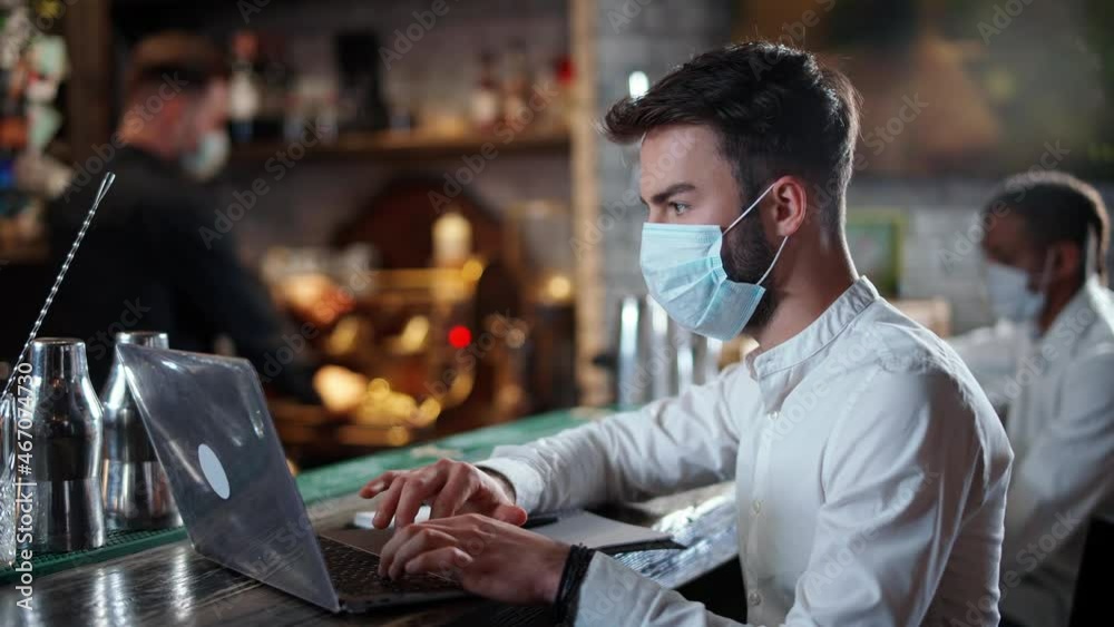Freelancer works outside the home, a young man developer in a medical mask sits at the bar and works using a laptop, working during a pandemic. Freelancer works outside the home, a young man developer in a medical mask sits at the bar and works using a laptop, working during a pandemic.