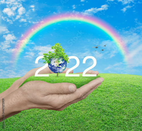2022 white text with planet and tree on green grass field in hands over blue sky, white clouds, and rainbow, Happy new year 2022 ecological cover, Save the earth concept, Elements of this image furnis