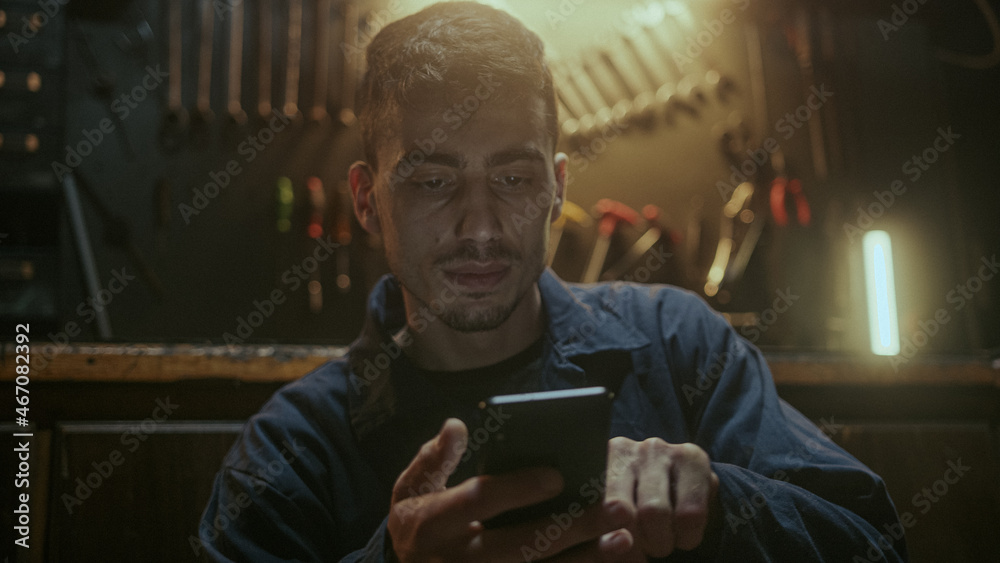 Young handsome blacksmith man sitting in front of the workbench, resting and using smartphone