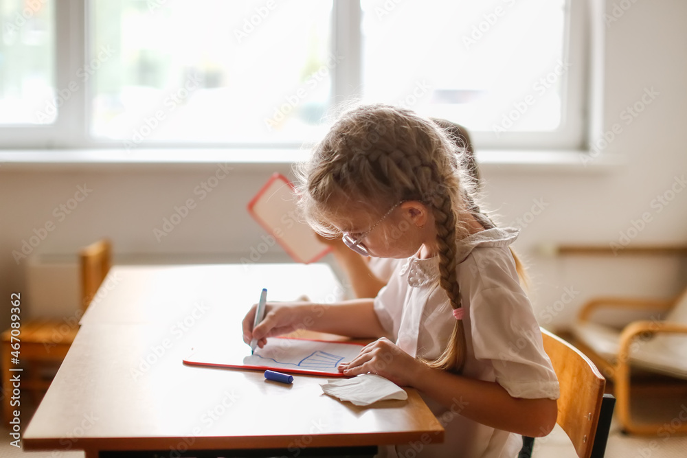 Cute caucasian girl child blonde with pigtails writes with a marker on a tablet, classes at school and independent work of children, light real class at school, soft focus and warm toning Cute caucasian girl child blonde with pigtails writes with a marker on a tablet, classes at school and independent work of children, light real class at school, soft focus and warm toning