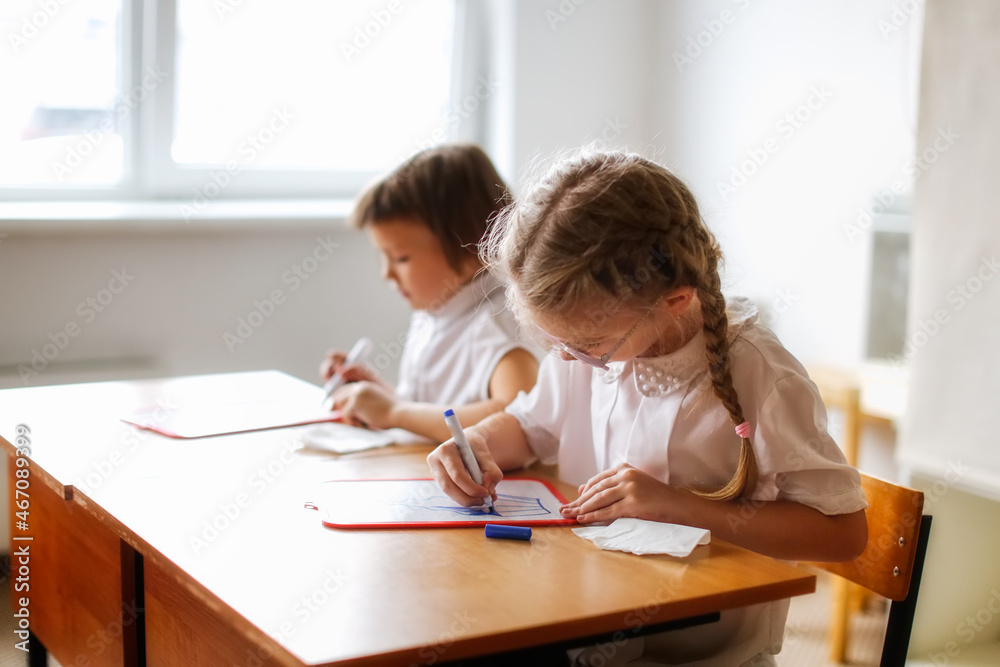 Cute caucasian girl child blonde with pigtails writes with a marker on a tablet, classes at school and independent work of children, light real class at school, soft focus and warm toning Cute caucasian girl child blonde with pigtails writes with a marker on a tablet, classes at school and independent work of children, light real class at school, soft focus and warm toning