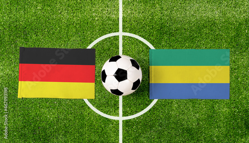 Top view soccer ball with Germany vs. Gabon flags match on green football field.