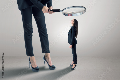Businessperson in suit using magnifier magnifying glass to look at tiny female subordinate on concrete background with shadow and mock up place. Hiring and dominance concept.