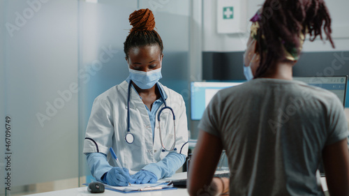 African american doctor preparing prescription paper for black patient at consultation during coronavirus pandemic. Medic and woman with face masks doing checkup for healing treatment.