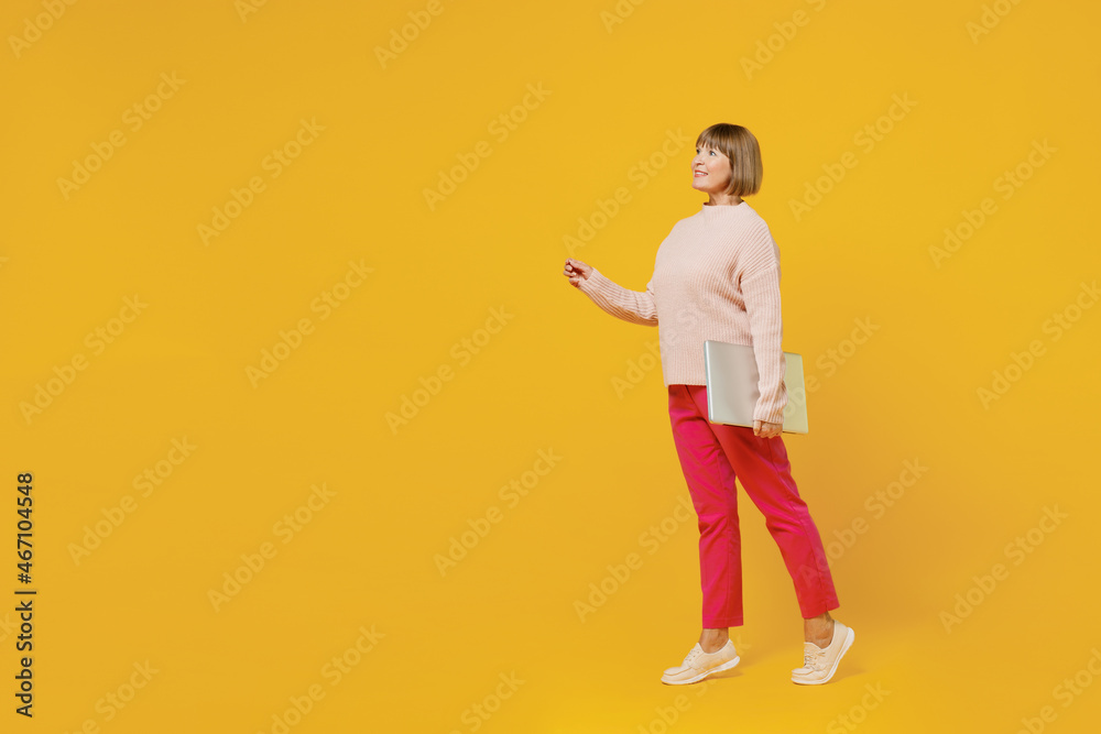 Full body elderly fun woman 50s wears pink casual knitted sweater hold in hand closed laptop pc computer walking go point finger aside on workspace isolated on plain yellow background studio portrait Full body elderly fun woman 50s wears pink casual knitted sweater hold in hand closed laptop pc computer walking go point finger aside on workspace isolated on plain yellow background studio portrait