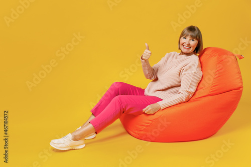 Full body smiling happy cheerful elderly caucasian woman 50s wear pink casual knitted sweater look camera show thumb up gesture sit in bag chair isolated on plain yellow background studio portrait