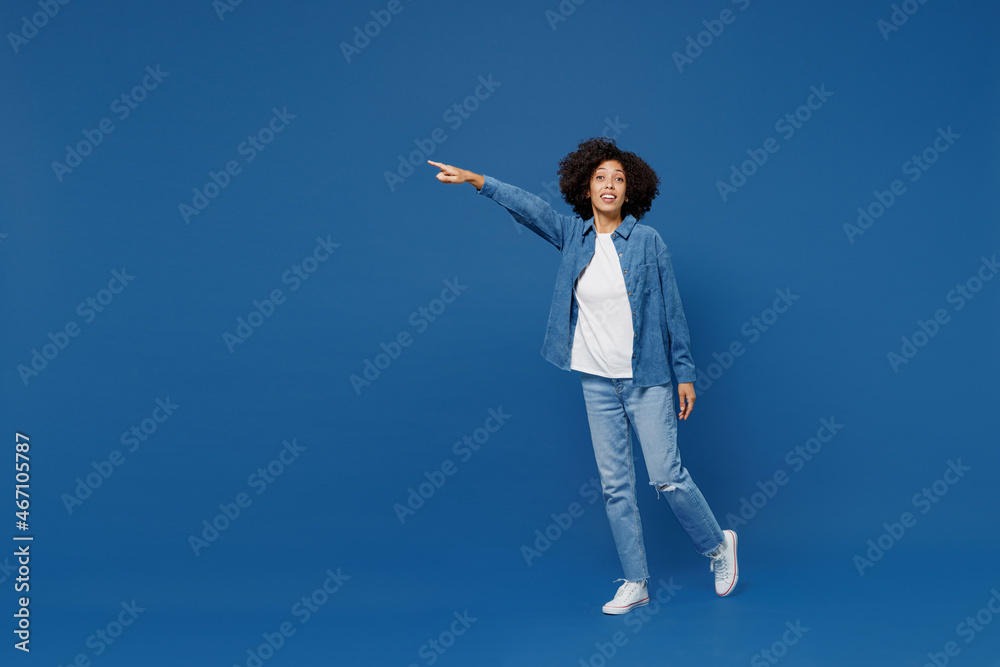 Full body side view young smiling happy black woman in casual clothes shirt white t-shirt walking going point index finger aside on workspace isolated on plain dark blue background studio portrait Full body side view young smiling happy black woman in casual clothes shirt white t-shirt walking going point index finger aside on workspace isolated on plain dark blue background studio portrait
