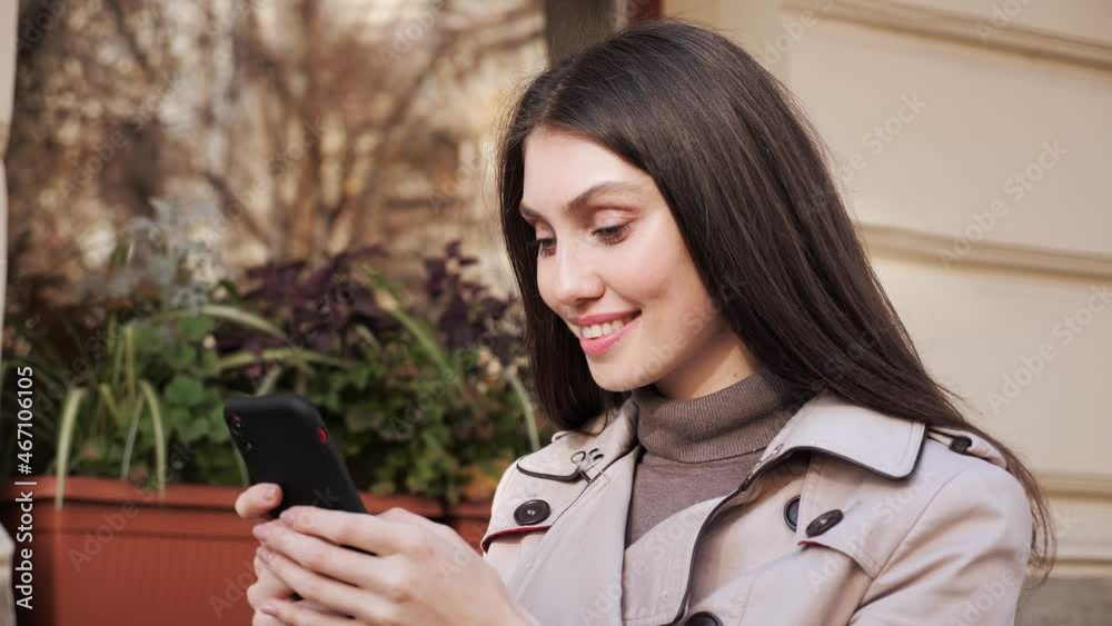 Portrait of happy beautiful Caucasian female girl texting on cellphone while standing on the terrace of the restaurant. Joyful young pretty girl in good mood typing tapping on smartphone outdoors. Portrait of happy beautiful Caucasian female girl texting on cellphone while standing on the terrace of the restaurant. Joyful young pretty girl in good mood typing tapping on smartphone outdoors.