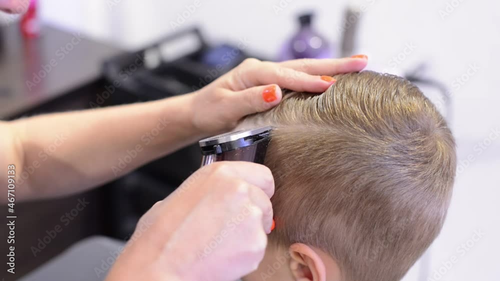 Hairdresser with a neat manicure cuts the hair of a child using professional trimmer while sitting in a barbershop close-up. Professional hairdresser cuts little blond boy with hair clipper. Hairdresser with a neat manicure cuts the hair of a child using professional trimmer while sitting in a barbershop close-up. Professional hairdresser cuts little blond boy with hair clipper.