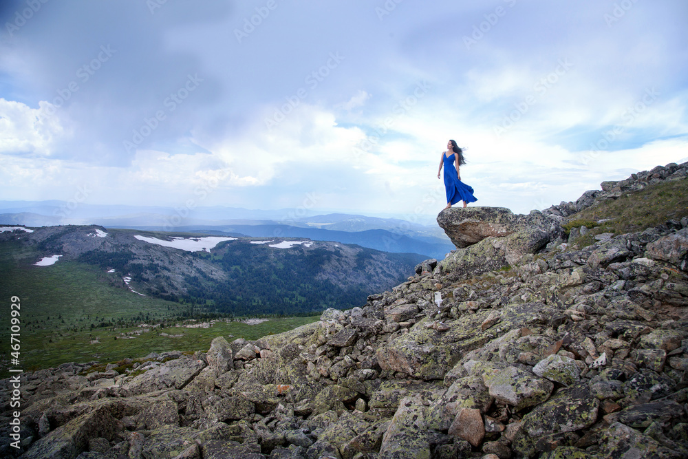 An Altai girl in a blue dress stands on top of the Sarlyk mountain above a cliff among rocks and stones. Tourism concept.