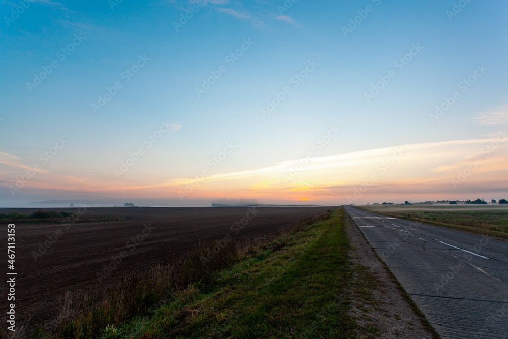 An empty asphalt road through the fields and forest in a thick fog at sunrise