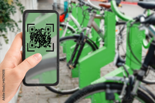 technology, transport and sustainability concept - close up of hand holding smartphone with qr code on screen over bicycles at electric bike charging station on city street on background