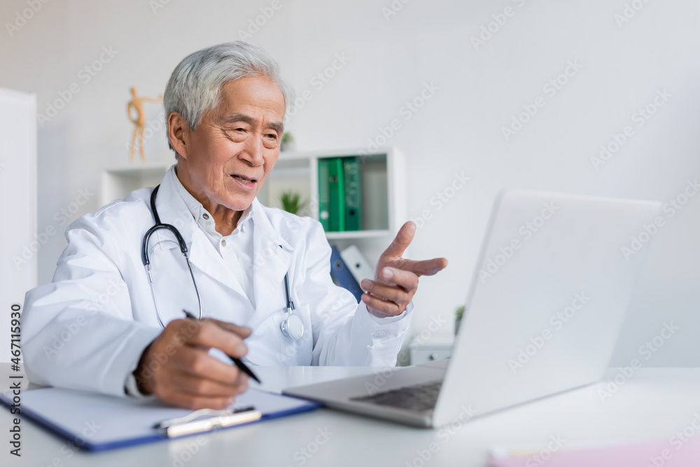 Elderly asian doctor pointing at laptop during video call and writing on clipboard in clinic Elderly asian doctor pointing at laptop during video call and writing on clipboard in clinic