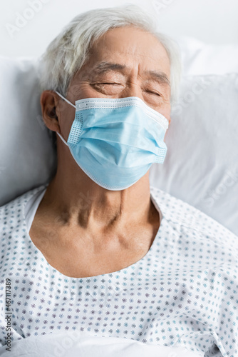 Senior asian patient in medical mask and gown lying on bed in clinic