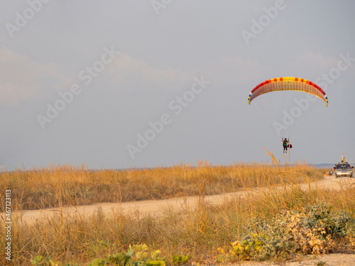 A landscape of yellow, sun-bleached grass in the steppe, over which a paraglider towers. The car pulls the cable and lifts the paraglider into the sky. A dirt road with overgrown grass along it