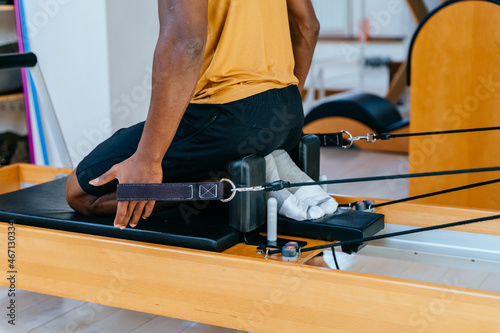 Hands of african fit man in sport clothing working out at gym. Flexible male pulling strings of a pilates reformer machine in fitness center, rowing. Close up. photo