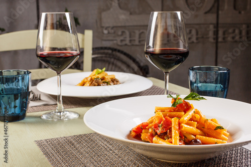 Tavolo di un ristorante apparecchiato con tovagliette, bicchieri per l'acqua, calici con vino rosso e due primi piatti di pasta