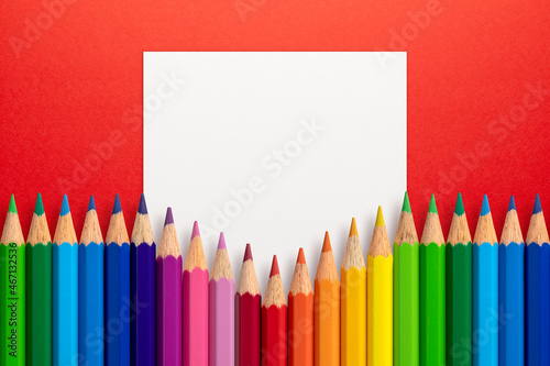 Creative layout made of color pencils on red paper background with white paper card note. Flat lay. Education concept.