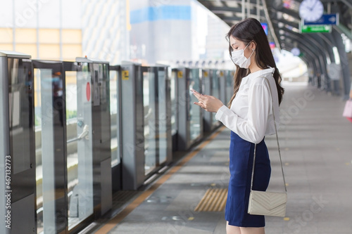 Asian professional business woman stands on a sky train station in town wearing a face mask while uses her smartphone to send messages  to the customer office © nut_foto