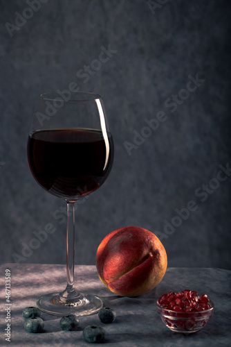 a simple still life. a beautiful glass of red wine, a glass plate with pomegranate seeds and a fresh peach on a dark background with creative lighting
