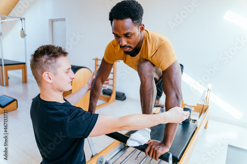 Two multiethnic men workout in modern light pilates studio interior. Fitness instructor corrects and controls the Pilates exercise that his african american male