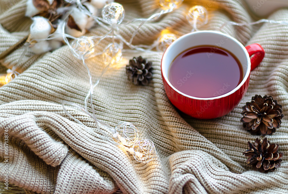 New Year's atmosphere. A red cup of tea, fir cones, a red satin ribbon with the inscription Merry Christmas and a glowing garland lying on a light knitted scarf New Year's atmosphere. A red cup of tea, fir cones, a red satin ribbon with the inscription Merry Christmas and a glowing garland lying on a light knitted scarf