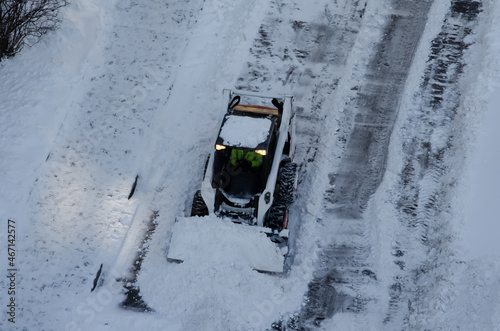 Skid steer loader removes snow from the city streets. Top view of the road with cars and snow blower. Seasonal work in winter snowy city. Equipment and city worker.