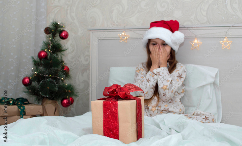 Christmas morning, a little girl in pajamas in bed rejoices with gifts on the background of a Christmas tree. A happy, smiling child opens a New Year's gift at home. The concept of holidays, Christmas