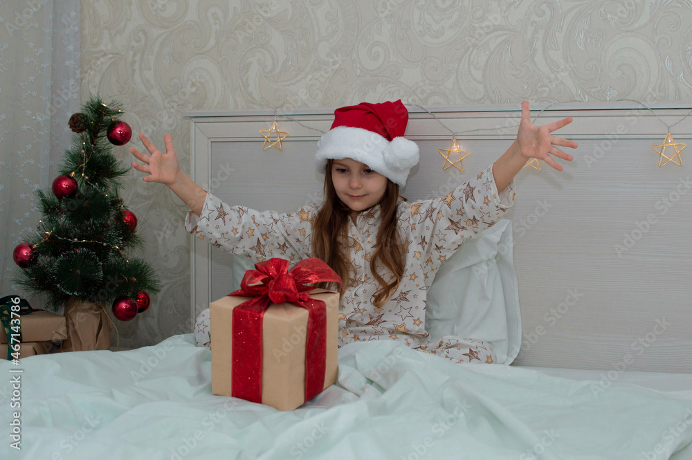 Christmas morning, a little girl in pajamas in bed rejoices with gifts on the background of a Christmas tree. A happy, smiling child opens a New Year's gift at home. The concept of holidays, Christmas