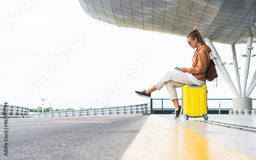 Young woman using digital tablet while sitting on wheeled luggage at airport photo