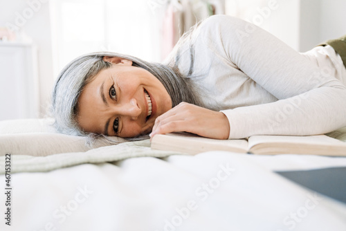 Grey asian woman smiling and reading book while lying on bed
