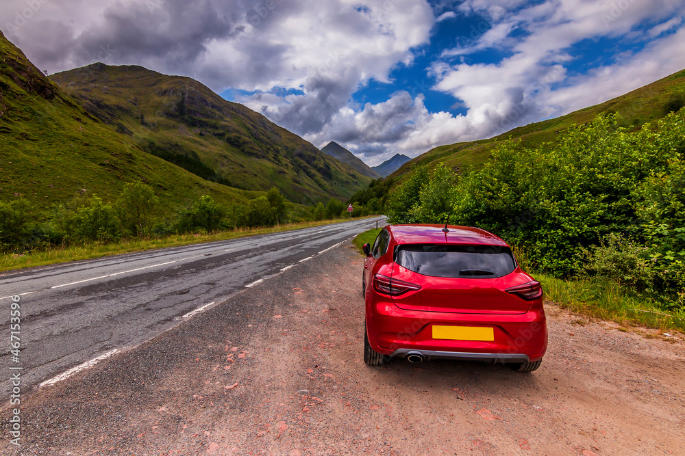 Naklejka premium Red car in the foreground next to a lonely road in sunshine. Green landscape with trees and bushes. Mountain range in the Scottish Highlands in summer with cloudy skies