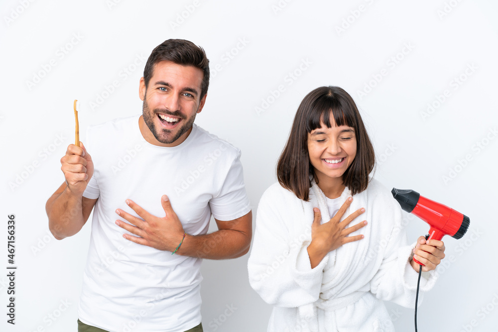 Young caucasian couple holding a hairdryer and toothbrush isolated on white background smiling a lot while putting hands on chest Young caucasian couple holding a hairdryer and toothbrush isolated on white background smiling a lot while putting hands on chest