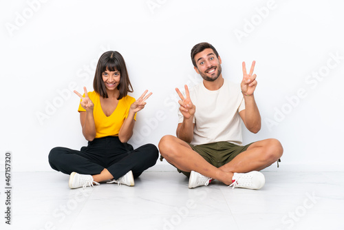 Young couple sitting on the floor isolated on white background smiling and showing victory sign with both hands