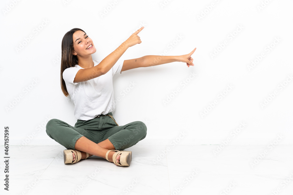 Teenager girl sitting on the floor pointing finger to the side and presenting a product