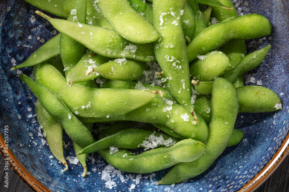 Organic steamed Japanese Edamame, a young soybean that is rich in nutrients served in blue bowl and sprinkled with sea salt flakes. Vegetables Background. Healthy eating concept Organic steamed Japanese Edamame, a young soybean that is rich in nutrients served in blue bowl and sprinkled with sea salt flakes. Vegetables Background. Healthy eating concept
